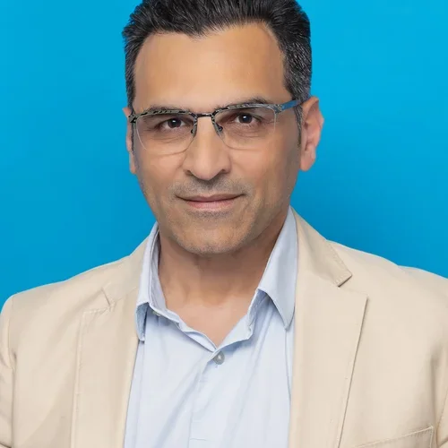 amir shaerzadeh wearing glasses and a cream colored suit on a bright blue background